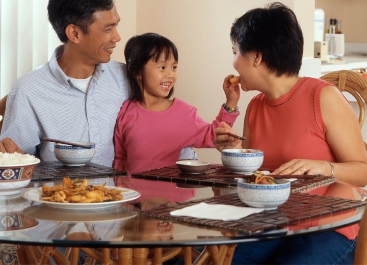 An Asian family, an adult male and female are seated around a table eating a meal with a young female standing in between the adults. Photographer Rhoda Baer
