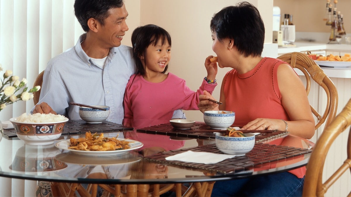 An Asian family, an adult male and female are seated around a table eating a meal with a young female standing in between the adults. Photographer Rhoda Baer